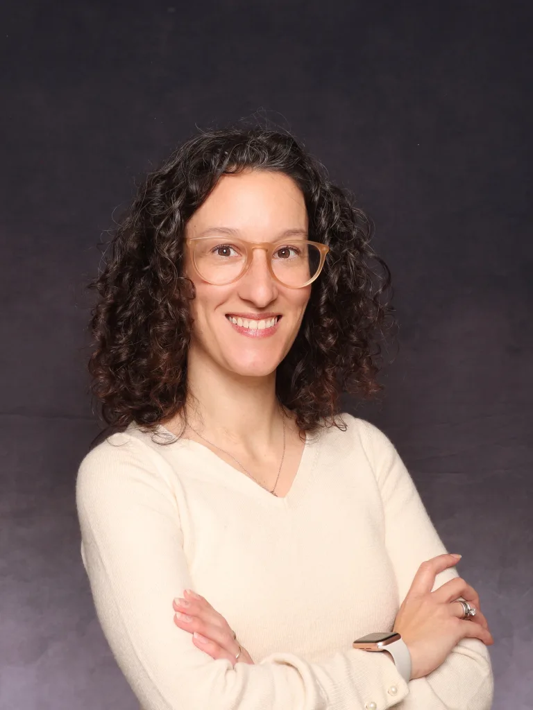 A person with curly brown hair and glasses, wearing a cream-colored sweater and a watch, smiling with arms crossed in front of a dark background.