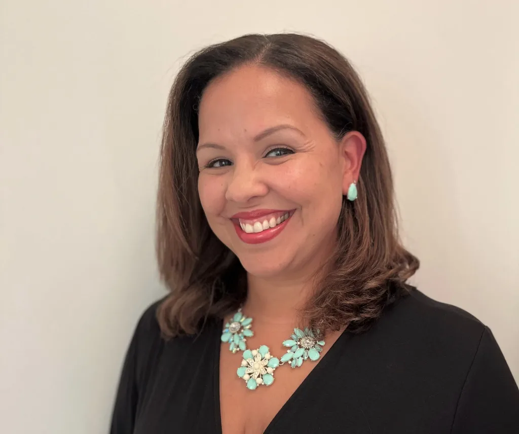 A woman with shoulder-length brown hair smiles at the camera. She wears a black top, turquoise earrings, and a matching floral necklace, standing against a plain light background.