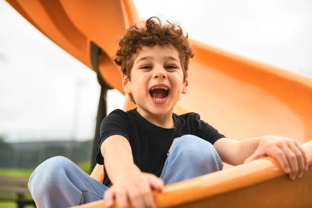 A young boy with curly hair, wearing a black shirt and light blue pants, smiles joyfully while sitting on an orange playground slide outdoors.