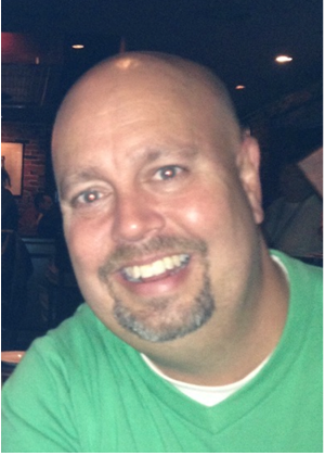 A man with a shaved head and a goatee, wearing a green shirt, smiles at the camera in a dimly lit indoor setting.