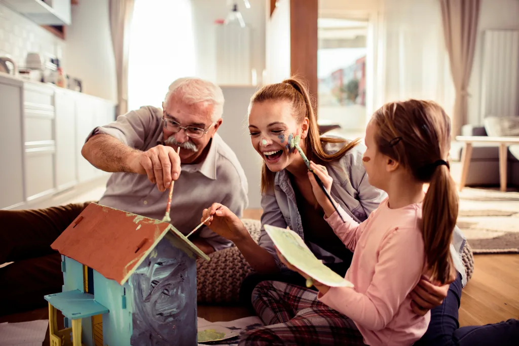 A cheerful family of three sits on the floor painting a birdhouse together, smiling and laughing as the woman playfully gets paint on her face. The room is bright and cozy, with sunlight streaming in.