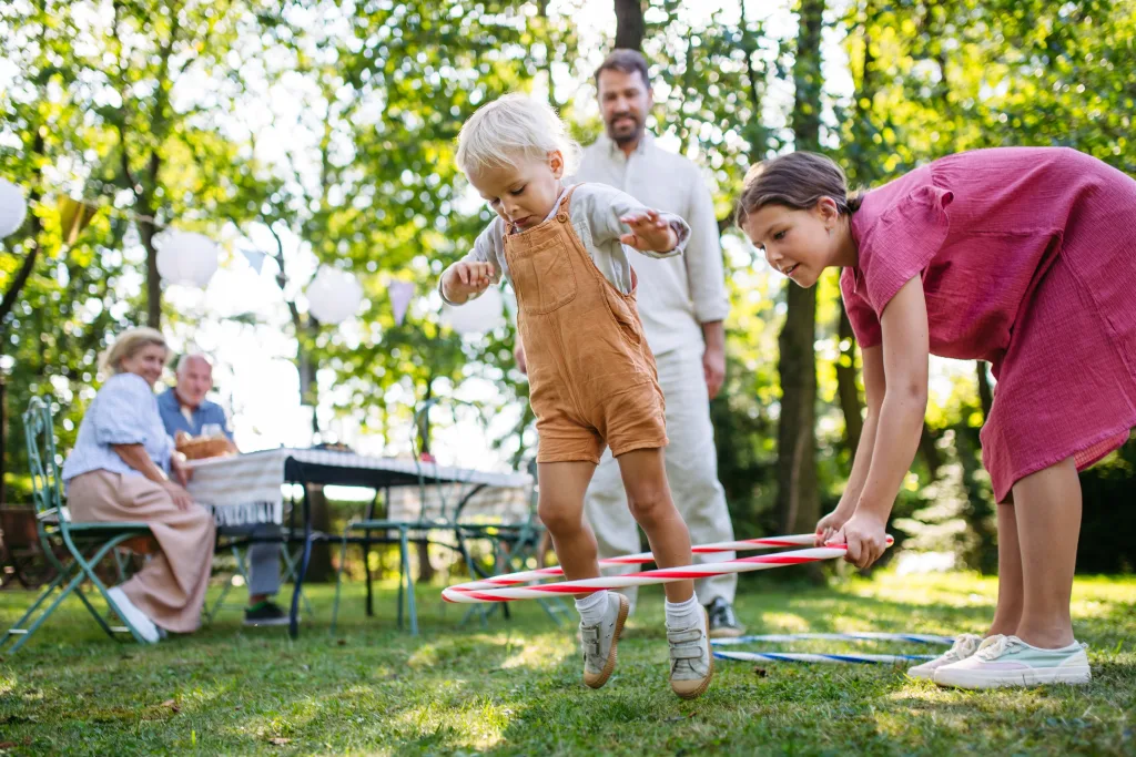 A young child jumps through a hula hoop held by a girl on a sunny day outdoors, while two adults sit at a table and another adult stands nearby in a grassy, wooded backyard.