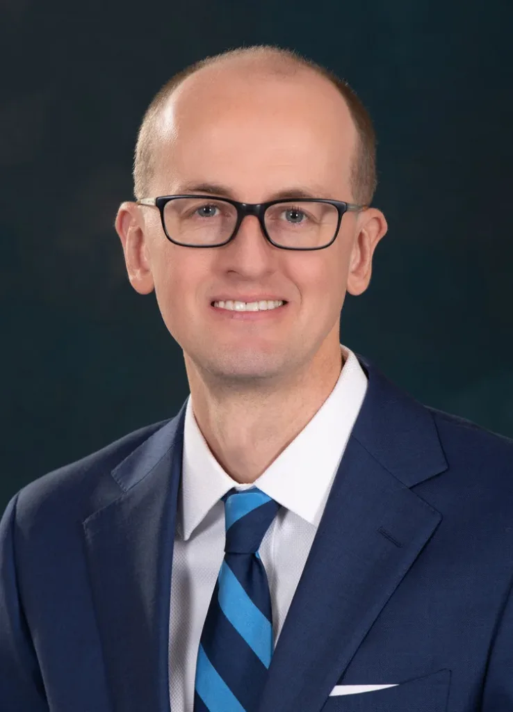 A man wearing glasses, a blue suit, white shirt, and blue striped tie, smiling in front of a dark background.