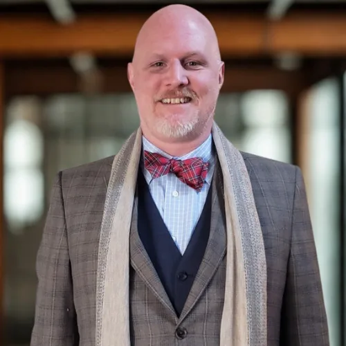 A smiling, bald man with a goatee wears a grey plaid suit, red bow tie, light blue shirt, and a beige scarf, standing indoors with blurred background.