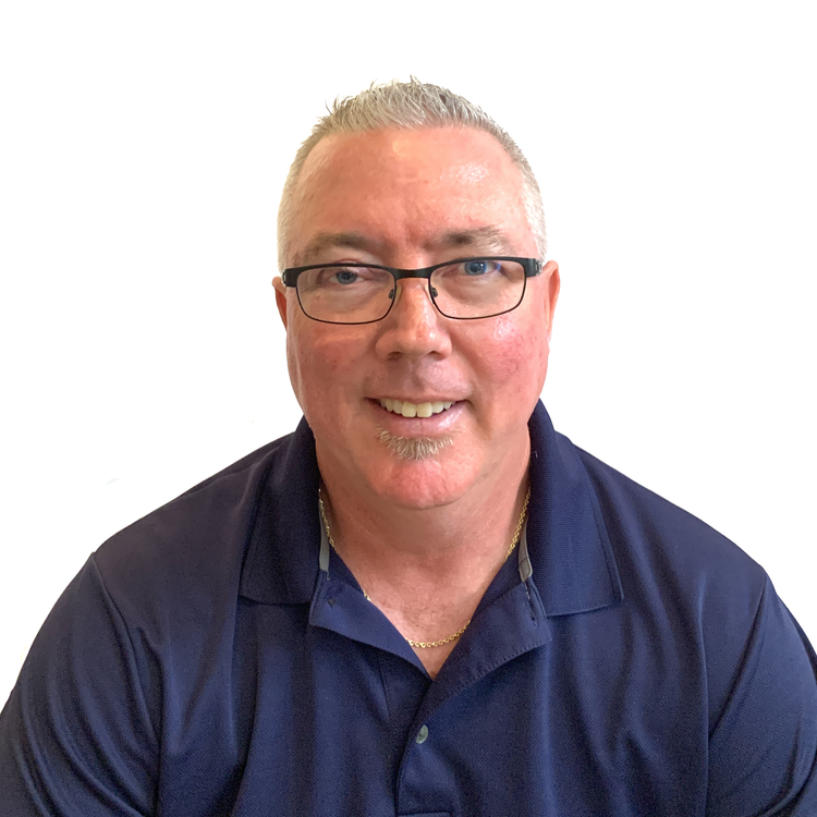 A middle-aged man with short gray hair, glasses, and a trimmed goatee smiles at the camera. He is wearing a navy blue collared shirt and a gold chain necklace. The background is plain white.