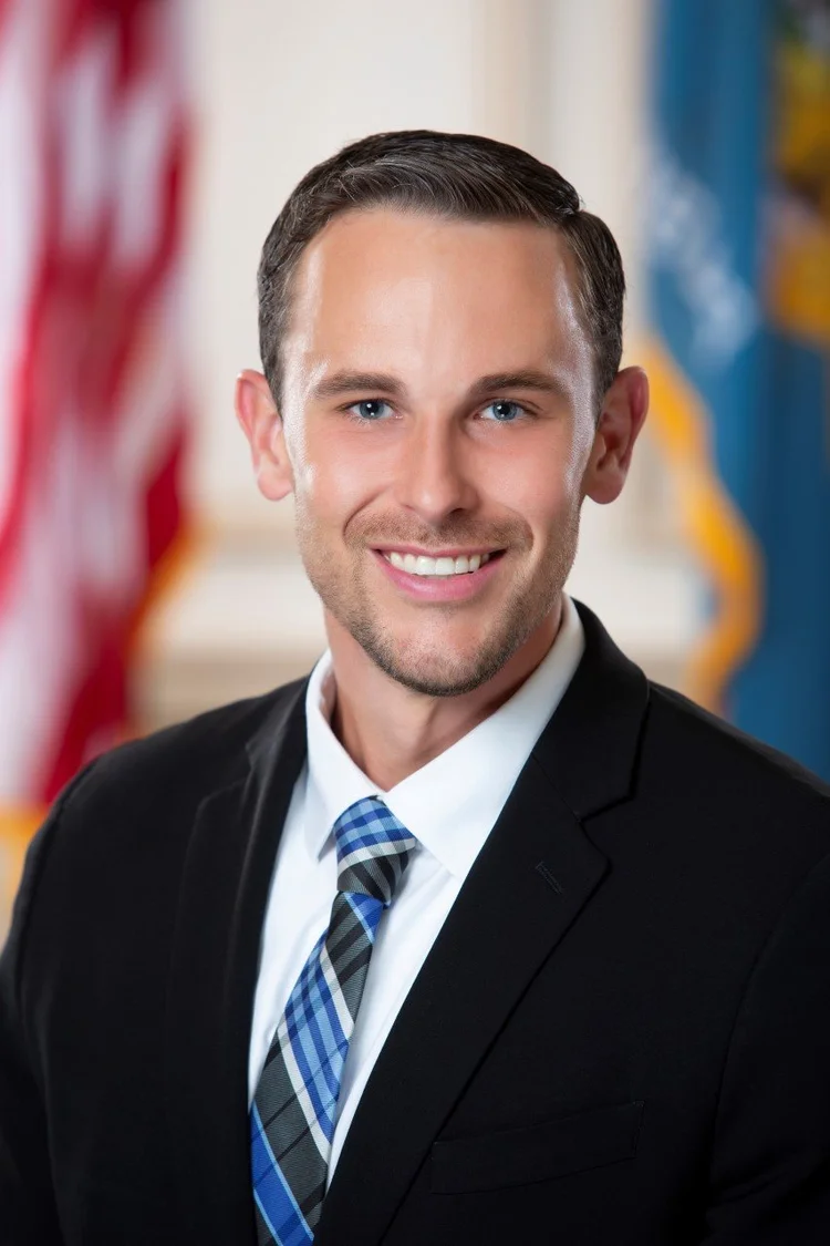 A man in a black suit, white shirt, and blue plaid tie smiles in front of blurred American and blue flags.