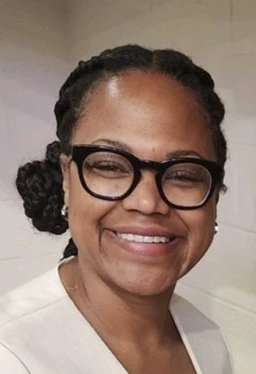 A woman with dark braided hair styled in a low bun, wearing black-rimmed glasses and a light-colored top, smiles at the camera against a neutral background.