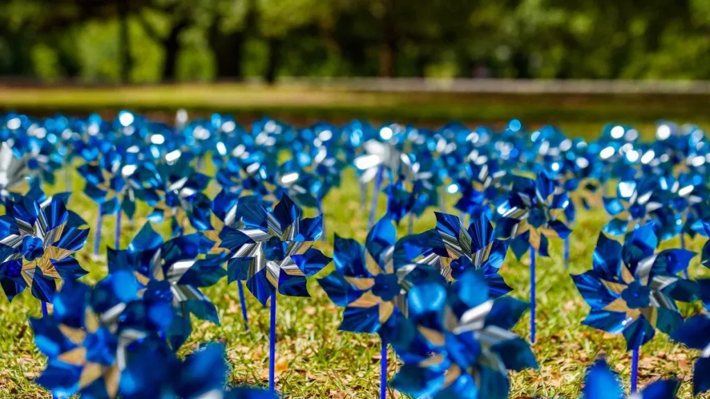 A field of shiny blue pinwheels is planted in grass, with trees and a blurred background, under bright daylight.