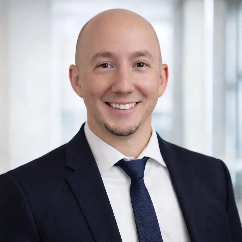 A smiling bald man in a dark suit, white shirt, and navy tie poses in front of a blurred office background.