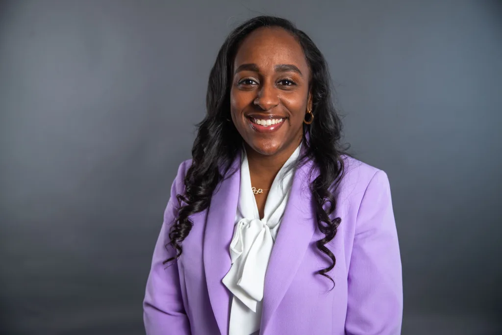 A woman with long dark hair smiles, wearing a light purple blazer over a white blouse, with a plain gray background.