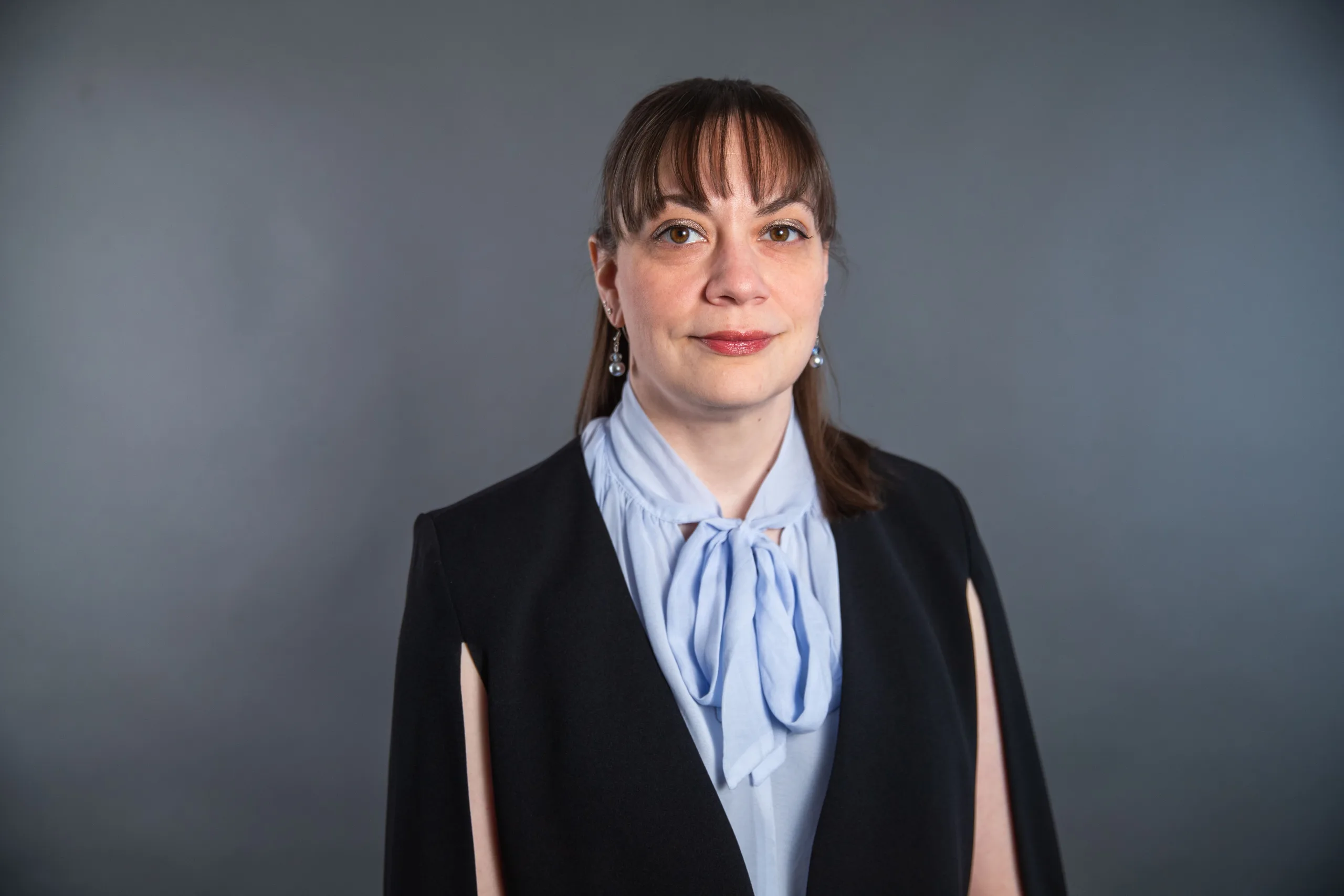 A woman with straight brown hair and bangs, wearing a black blazer and a light blue blouse with a bow, stands in front of a plain gray background, looking at the camera with a slight smile.