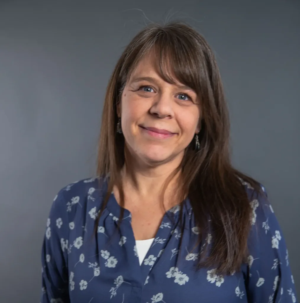 A woman with long brown hair wearing a blue blouse with a white floral pattern stands in front of a plain gray background, smiling gently at the camera.