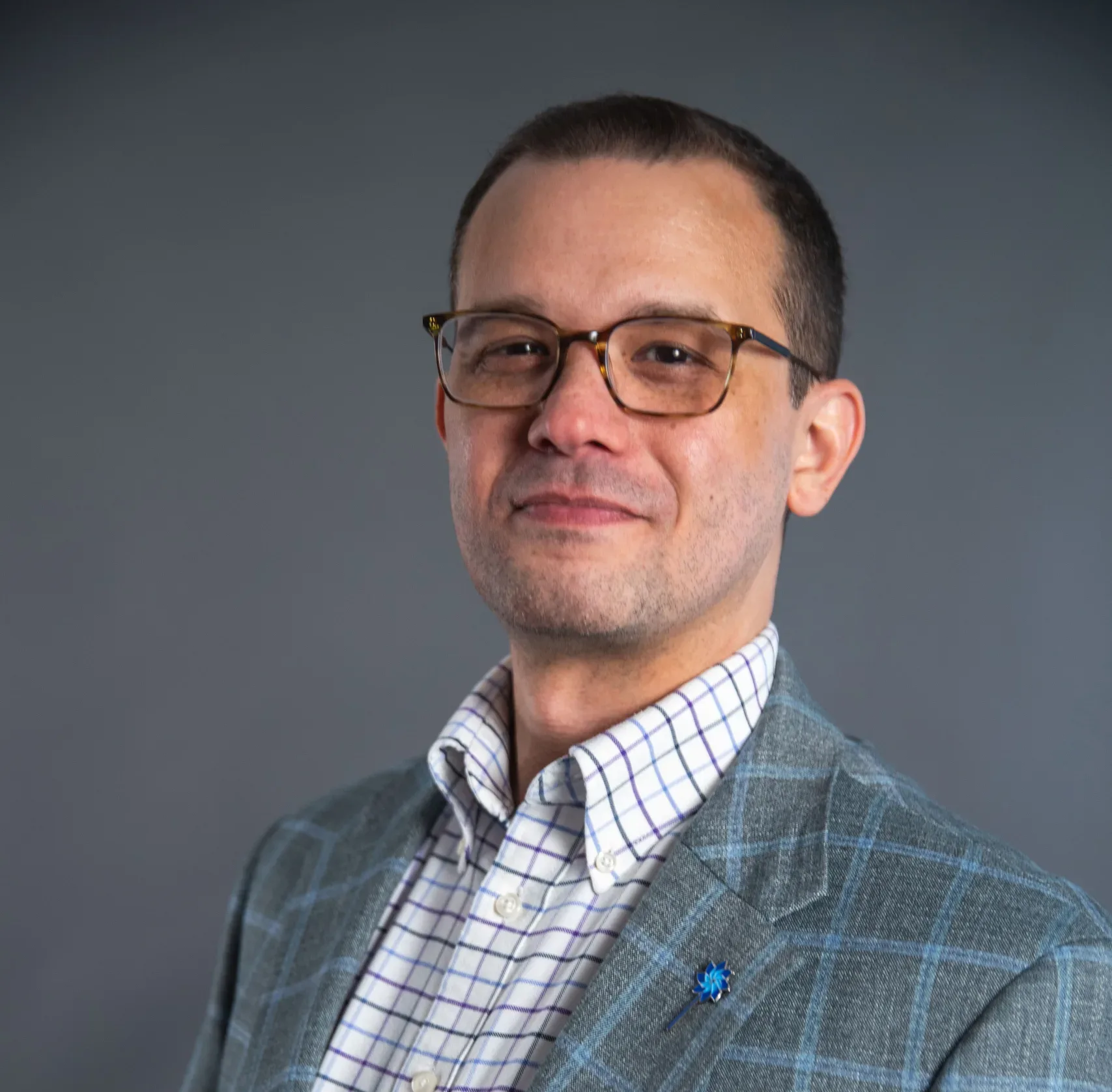 A man wearing glasses, a checked dress shirt, and a gray plaid suit jacket stands in front of a plain gray background, smiling slightly at the camera.