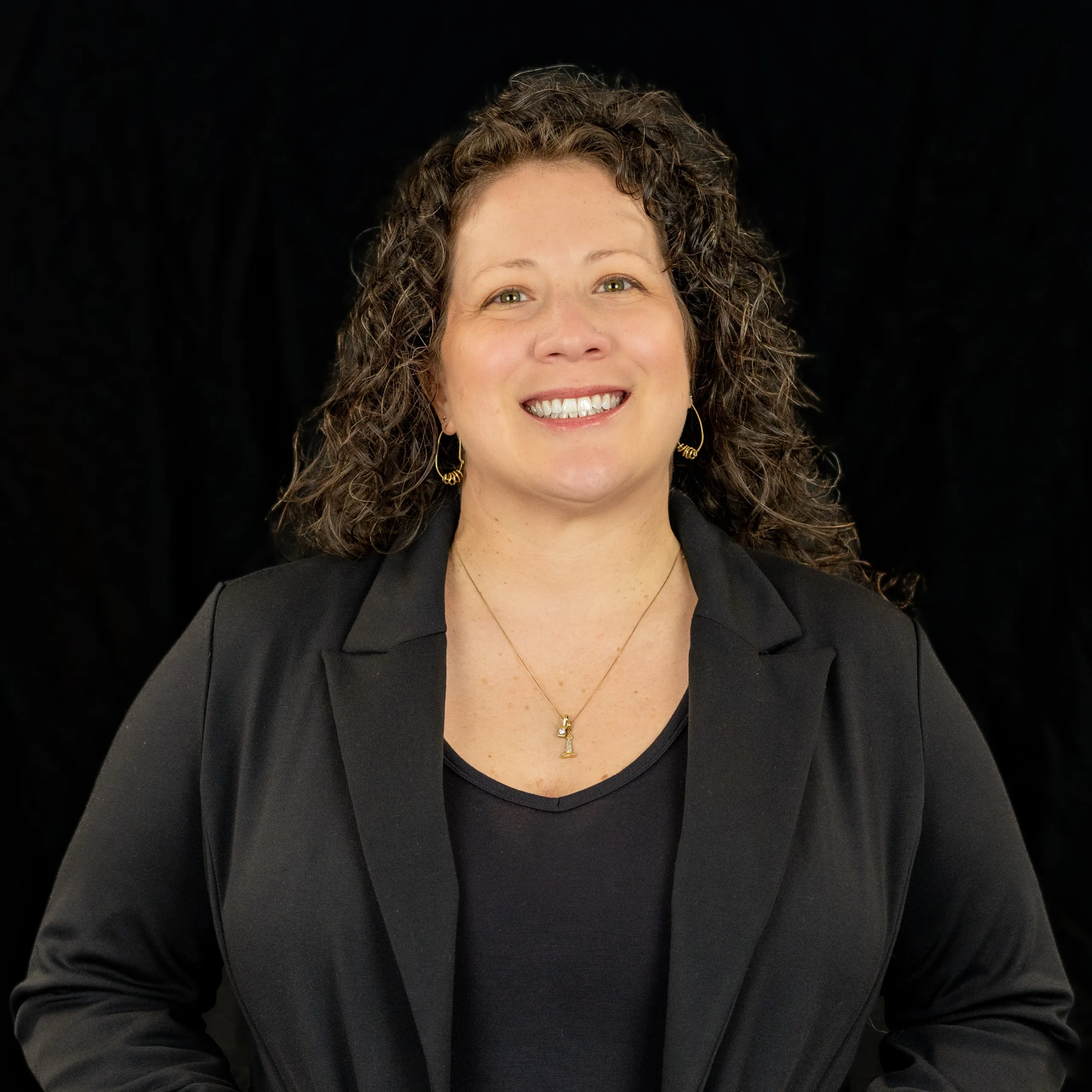 A woman with curly brown hair, wearing a black blazer, black top, and gold necklace, smiles confidently against a plain black background.