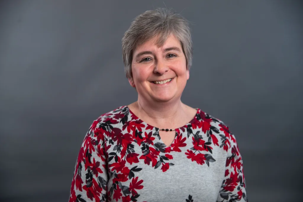 A smiling woman with short gray hair wears a gray sweater with a red and black floral pattern, standing in front of a plain dark gray background.