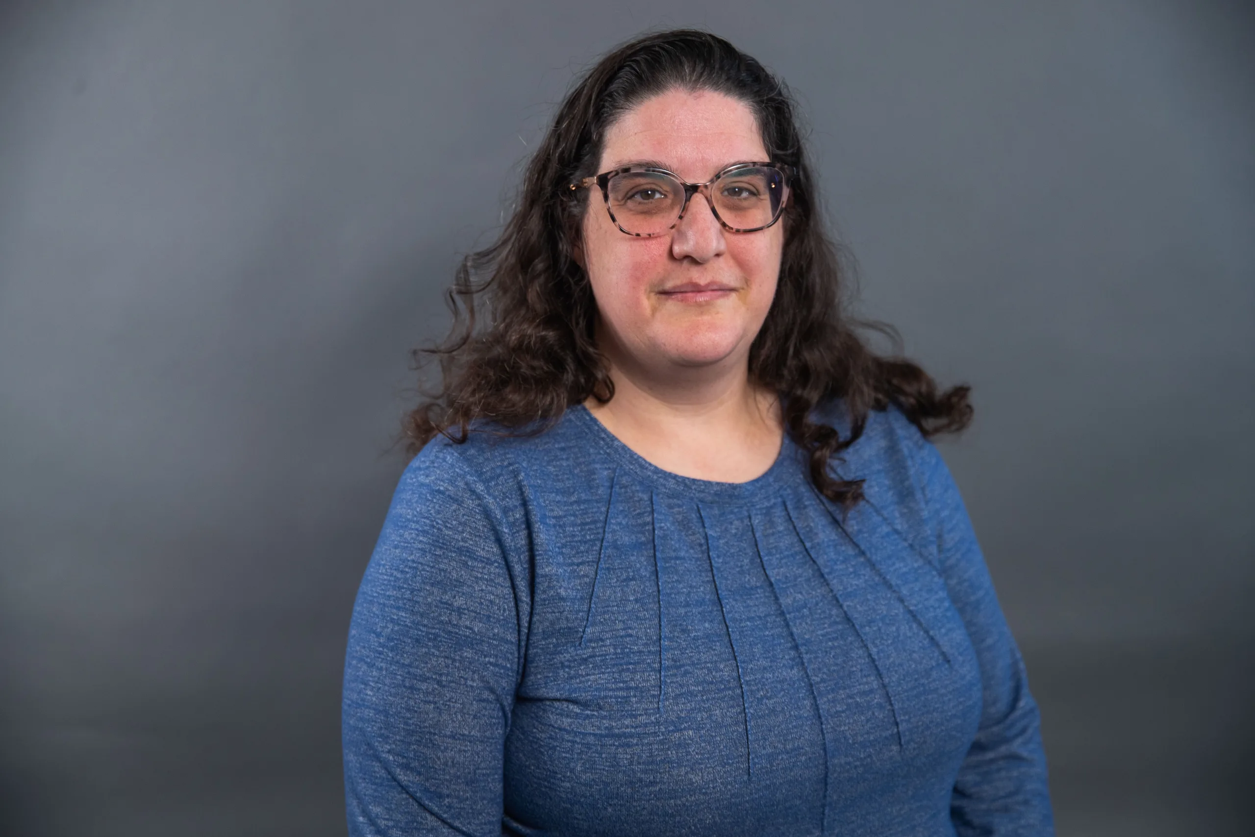 A woman with long, wavy brown hair and glasses is wearing a blue, long-sleeve top. She is standing in front of a plain gray background, facing the camera with a neutral expression.