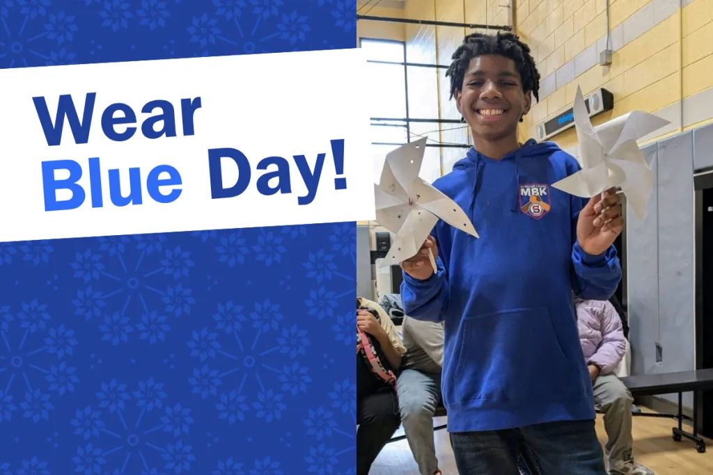 A smiling student in a blue hoodie holds two white paper pinwheels. Behind him are people seated in a gym. The left side of the image has a floral blue background with text reading Wear Blue Day!.